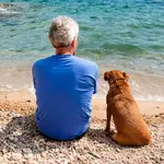 An old man and his dog sitting on a beach looking out at the water