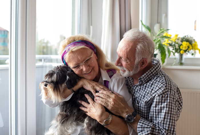 An older couple is hugging a small dog.