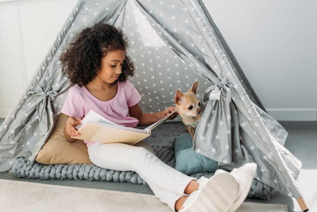 A girl is sitting in a tent with her Chihuahua, reading a book.